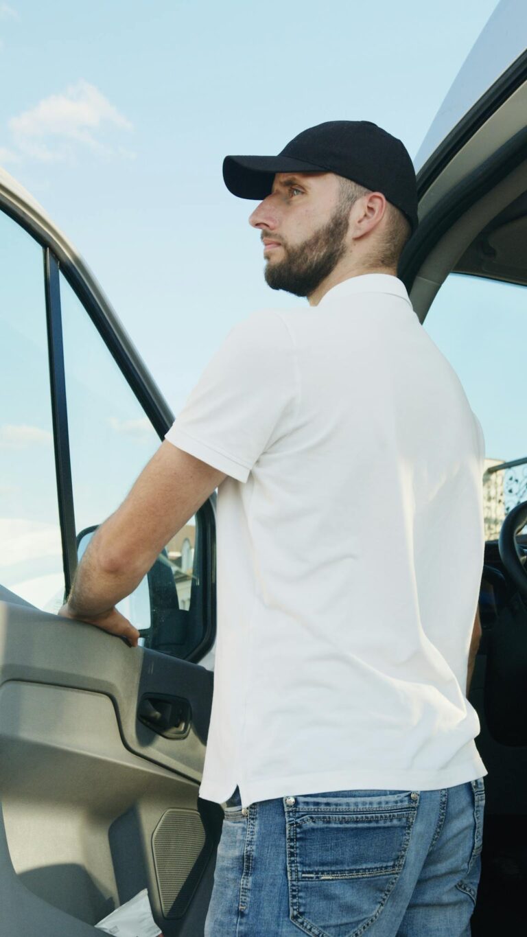 A person wearing a cap stands beside an open van door under a clear sky.