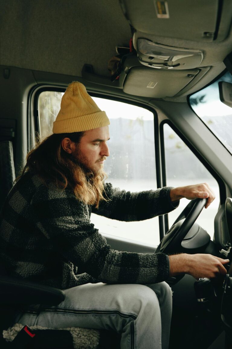 Side view of a man driving a van, wearing a yellow beanie, with a focused expression.